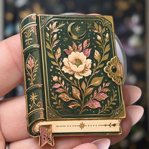 May include: A rectangular enamel pin shaped like a vintage book with a dark green cover, gold metallic accents, and a central floral illustration. The design features a crescent moon, leafy vines, a decorative clasp, and a small bookmark hanging below.