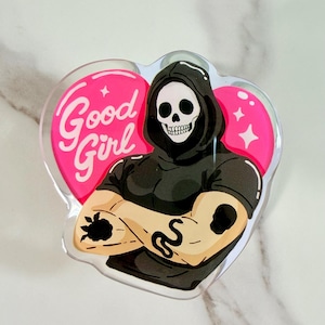 May include: A clear acrylic pin featuring a hooded skeleton figure with crossed arms, wearing a black shirt. The figure has tattoos on both arms. Behind the figure is a bright pink heart shape with the text Good Girl and two small white stars.