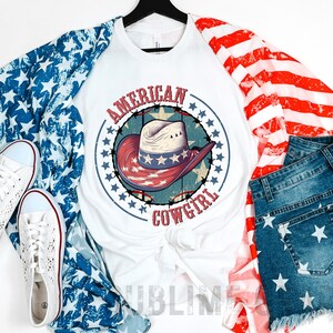 May include: White t-shirt with a red, white, and blue cowboy hat graphic and the text "AMERICAN COWGIRL". The shirt is layered over a blue and white star patterned fabric and a red and white striped fabric. A pair of white sneakers and blue denim shorts with white stars are also pictured.