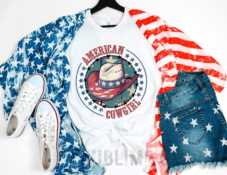 May include: White t-shirt with a red, white, and blue cowboy hat graphic and the text "AMERICAN COWGIRL". The shirt is layered over a blue and white star patterned fabric and a red and white striped fabric. A pair of white sneakers and blue denim shorts with white stars are also pictured.