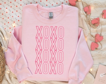 May include: A pink sweatshirt with the word "XOXO" repeated four times in a vertical line down the center of the shirt. The letters are outlined in a darker pink.