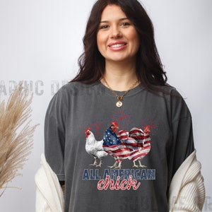 May include: Dark gray t-shirt featuring three chickens. One chicken is white, and the other two are designed with the American flag. The text "ALL AMERICAN chick" is printed below the chickens in blue and red.