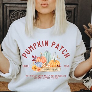 May include: Cream-colored sweatshirt with a "Pumpkin Patch" graphic. The design includes colorful pumpkins, leaves, and the text "Farm Fresh" and "Since 1953". Additional text includes "Hay Rides, Corn Maze, Hot Chocolate, Caramel Apples, Cider".