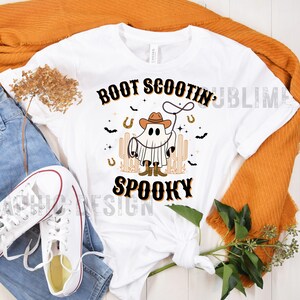 May include: White t-shirt with a black and orange graphic of a ghost wearing a cowboy hat and boots, holding a lasso. The text "BOOT SCOOTIN' SPOOKY" is printed in black and orange.