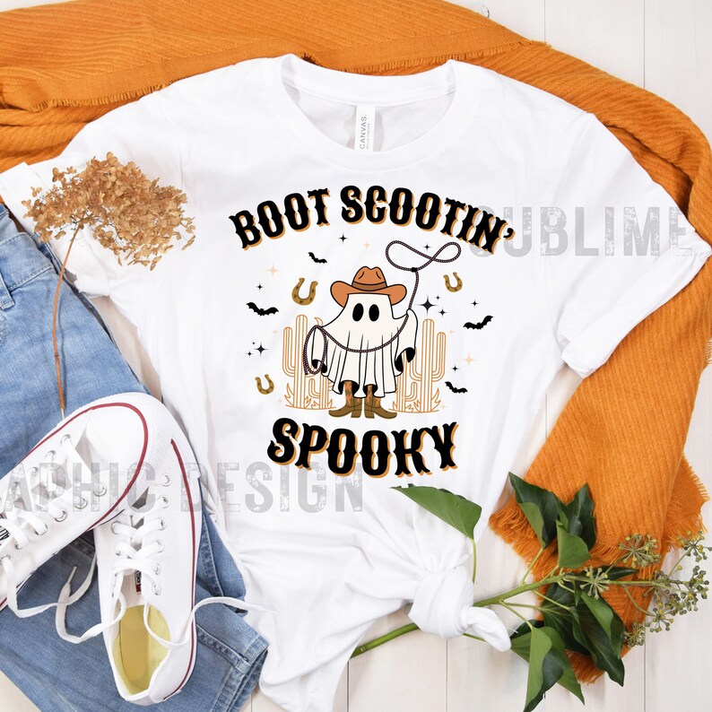 May include: White t-shirt with a black and orange graphic of a ghost wearing a cowboy hat and boots, holding a lasso. The text "BOOT SCOOTIN' SPOOKY" is printed in black and orange.