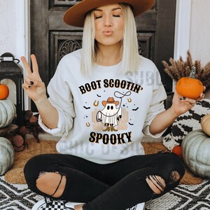 May include: A white sweatshirt with a graphic of a ghost wearing a cowboy hat and boots. The text "Boot Scootin' Spooky" is printed on the sweatshirt.