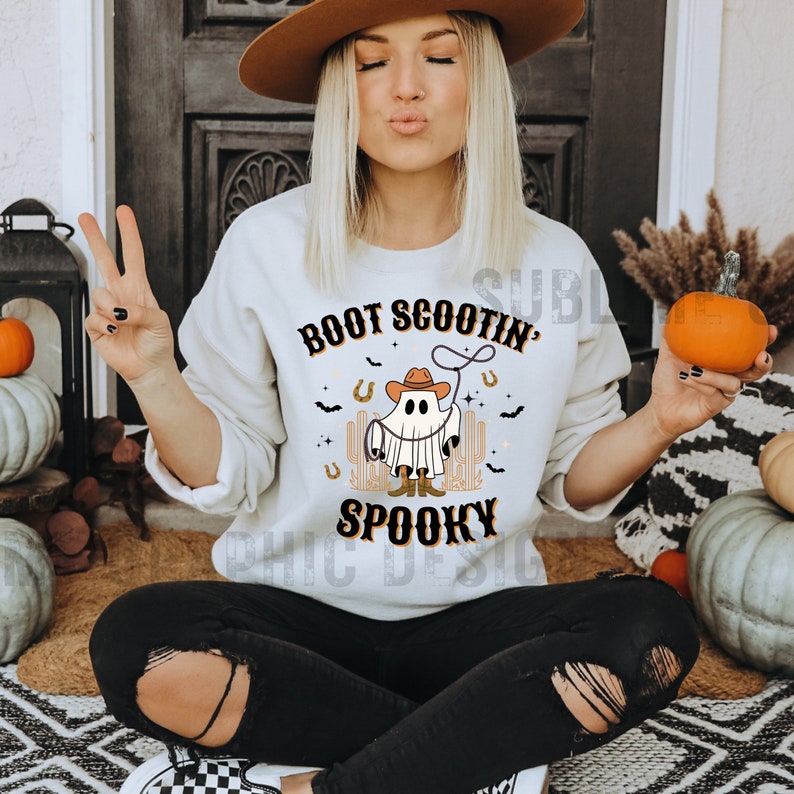 May include: A white sweatshirt with a graphic of a ghost wearing a cowboy hat and boots. The text "Boot Scootin' Spooky" is printed on the sweatshirt.