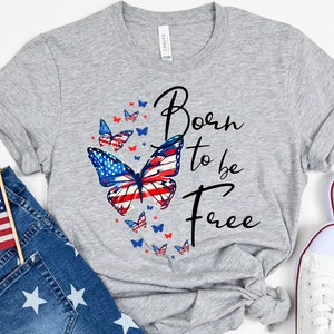 May include: Gray t-shirt with a red, white, and blue butterfly design. The butterfly has the American flag pattern. The text "Born to be Free" is printed in black below the butterfly.