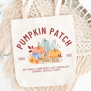 May include: Cream-colored tote bag with the words "Pumpkin Patch" in a burgundy font. The design includes watercolor pumpkins in orange, blue, and green, with the text "Farm Fresh" and "Since 1953". Additional text includes "Hay Rides, Corn Maze, Hot Chocolate, Caramel Apples, Cider".