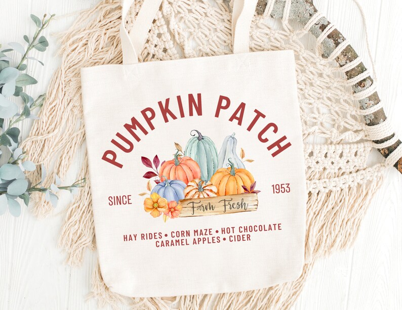 May include: Cream-colored tote bag with the words "Pumpkin Patch" in a burgundy font. The design includes watercolor pumpkins in orange, blue, and green, with the text "Farm Fresh" and "Since 1953". Additional text includes "Hay Rides, Corn Maze, Hot Chocolate, Caramel Apples, Cider".