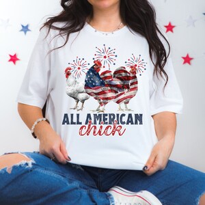 May include: White t-shirt featuring three rooster illustrations with patriotic American flag designs. The text "ALL AMERICAN chick" is printed below the rooster illustrations. Red, white, and blue fireworks are above the rooster illustrations.
