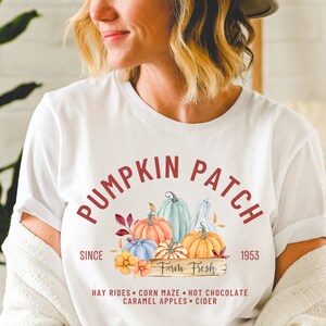 May include: White t-shirt with a "Pumpkin Patch" design in burgundy. The design features watercolor pumpkins in orange, blue, and green, with the text "Farm Fresh" and "Since 1953". Additional text includes "Hay Rides, Corn Maze, Hot Chocolate, Caramel Apples, Cider".