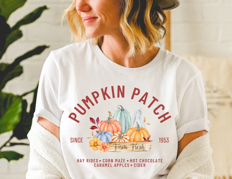 May include: White t-shirt with a "Pumpkin Patch" design in burgundy. The design features watercolor pumpkins in orange, blue, and green, with the text "Farm Fresh" and "Since 1953". Additional text includes "Hay Rides, Corn Maze, Hot Chocolate, Caramel Apples, Cider".