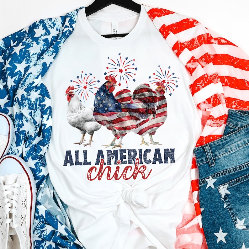May include: White t-shirt with raglan sleeves featuring a patriotic design. The shirt displays three roosters, one white and two with an American flag design, and fireworks. The text "ALL AMERICAN chick" is printed below the roosters.