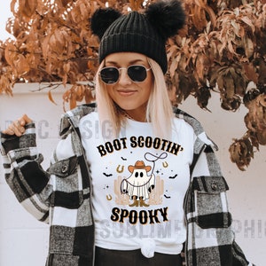May include: White t-shirt with a graphic of a ghost wearing a cowboy hat and boots, holding a lasso. The text "Boot Scootin' Spooky" is printed on the shirt.