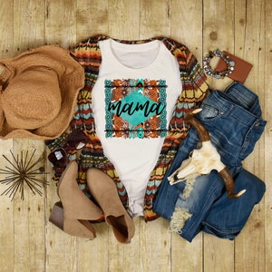 May include: White t-shirt with a turquoise and orange floral design and the word "mama" in a white font. The shirt is styled with a brown hat, a brown and white cardigan, blue jeans, and brown boots.