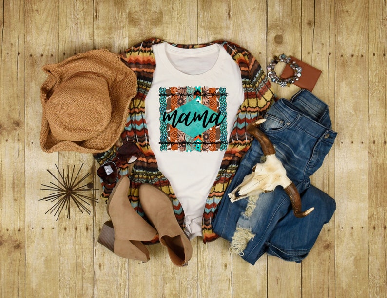 May include: White t-shirt with a turquoise and orange floral design and the word "mama" in a white font. The shirt is styled with a brown hat, a brown and white cardigan, blue jeans, and brown boots.