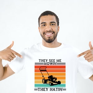 May include: A white t-shirt with a retro-style graphic of a lawnmower. The text on the shirt reads "They See Me Mowin' They Hatin'".