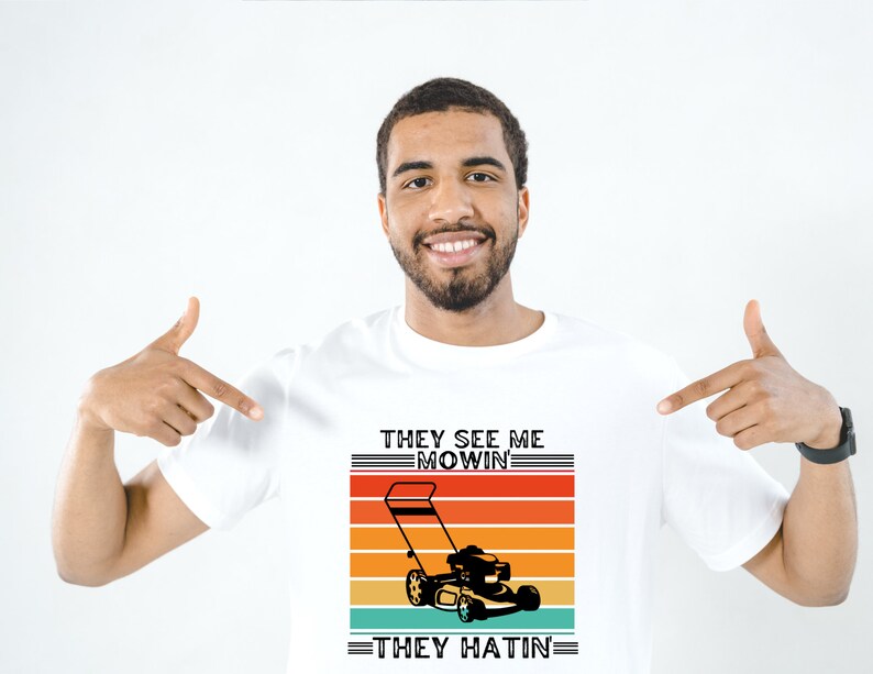 May include: A white t-shirt with a retro-style graphic of a lawnmower. The text on the shirt reads "They See Me Mowin' They Hatin'".