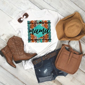 May include: White t-shirt with a turquoise and brown graphic design that says "mama" in a diamond shape. The shirt is styled with brown cowboy boots, a brown hat, a brown leather bag, and blue denim shorts.