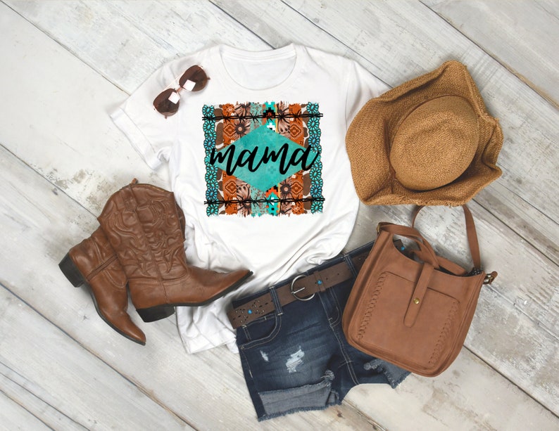 May include: White t-shirt with a turquoise and brown graphic design that says "mama" in a diamond shape. The shirt is styled with brown cowboy boots, a brown hat, a brown leather bag, and blue denim shorts.
