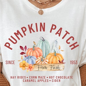 May include: White t-shirt with a fall-themed graphic. The design features the words "Pumpkin Patch" in a semi-circle, with illustrations of pumpkins, leaves, and flowers. The text "Farm Fresh" is also included, along with other fall activities.