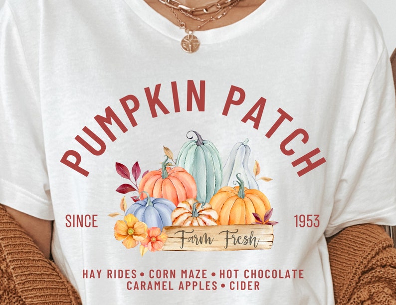 May include: White t-shirt with a fall-themed graphic. The design features the words "Pumpkin Patch" in a semi-circle, with illustrations of pumpkins, leaves, and flowers. The text "Farm Fresh" is also included, along with other fall activities.