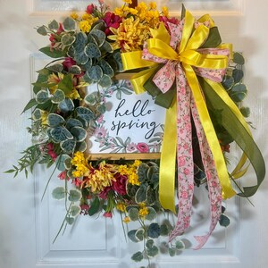 May include: A spring wreath with a wooden frame, adorned with colorful flowers, greenery, and a large yellow bow with pink floral ribbon. The frame displays the text "hello spring".