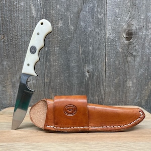May include: A white handled knife with a leather sheath. The sheath has the text "Moore Maker Inc. Chattanooga TN" embossed on it.