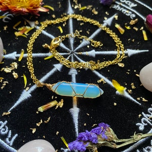 May include: A gold chain necklace with a light blue opal pendant wrapped in gold wire. The necklace is laying on a black velvet surface with gold leaf accents and dried flowers.