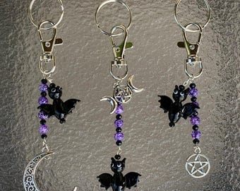 May include: Three silver keychains with purple and black beaded charms. Each keychain features a black bat charm, a silver crescent moon charm, a silver pentagram charm, and a silver triple moon charm.