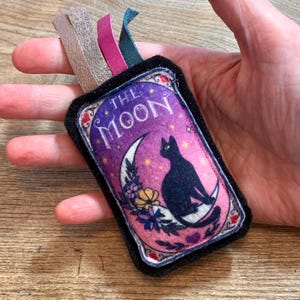 May include: A black felt pouch with a colorful image of a black cat sitting on a crescent moon. The image is surrounded by stars and flowers. The text "THE MOON" is printed in white on a purple background.
