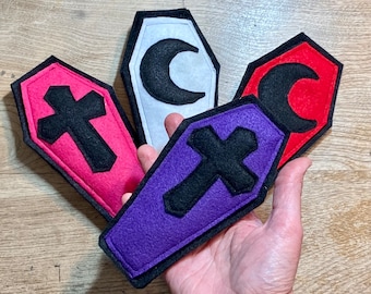 May include: Four felt coffin-shaped ornaments with different designs. One is white with a black crescent moon, one is red with a black crescent moon, one is pink with a black cross, and one is purple with a black cross.