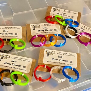 Pode incluir: Tr&ecirc;s conjuntos de tr&ecirc;s an&eacute;is de pl&aacute;stico coloridos impressos em 3D. Cada conjunto &eacute; etiquetado como "3pk Ring Things" e inclui um endere&ccedil;o de site. Os an&eacute;is s&atilde;o de v&aacute;rias cores, incluindo vermelho, laranja, amarelo, verde, azul, roxo, preto e branco.