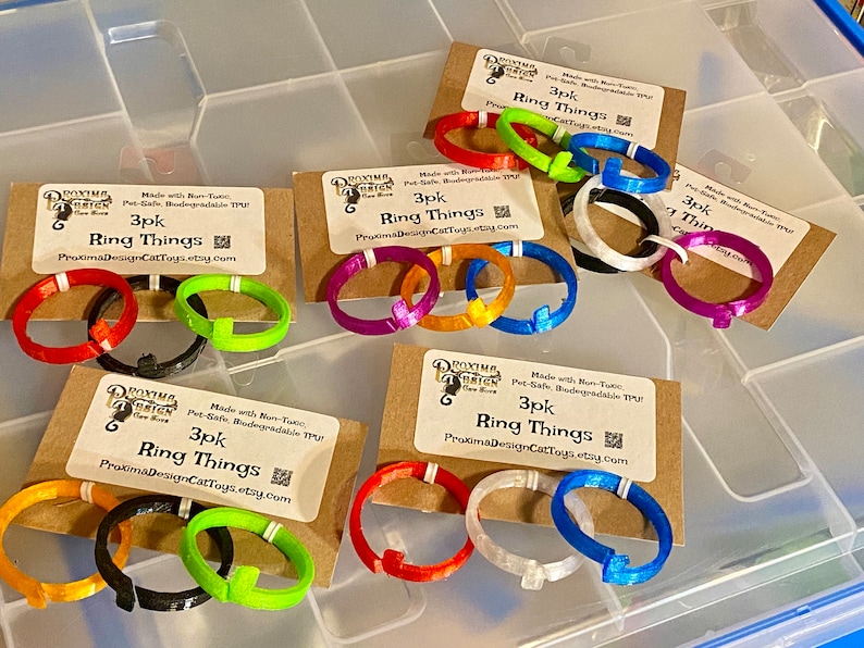 Pode incluir: Tr&ecirc;s conjuntos de tr&ecirc;s an&eacute;is de pl&aacute;stico coloridos impressos em 3D. Cada conjunto &eacute; etiquetado como "3pk Ring Things" e inclui um endere&ccedil;o de site. Os an&eacute;is s&atilde;o de v&aacute;rias cores, incluindo vermelho, laranja, amarelo, verde, azul, roxo, preto e branco.