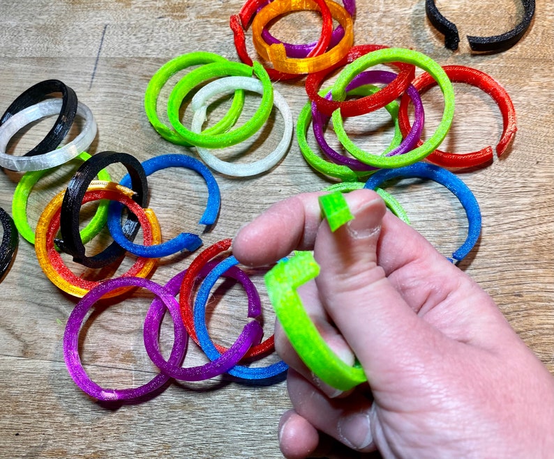 Pode incluir: Uma cole&ccedil;&atilde;o de an&eacute;is de pl&aacute;stico coloridos em v&aacute;rios tamanhos e cores, incluindo verde, azul, vermelho, laranja, roxo, preto e branco. Os an&eacute;is est&atilde;o dispostos em um padr&atilde;o aleat&oacute;rio sobre uma superf&iacute;cie de madeira.
