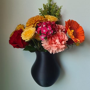 May include: A black ceramic vase with a variety of colorful flowers, including red, orange, yellow, pink, and white. The vase is mounted on a wall.