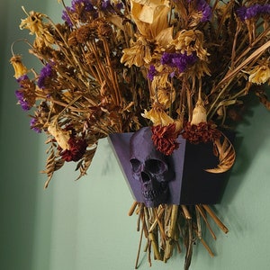May include: A dried flower arrangement in a black skull-shaped vase. The vase is made of a dark purple material and has a three-dimensional skull design. The flowers are a mix of dried purple, yellow, and brown flowers.