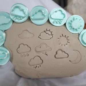Puede incluir: Un conjunto de 10 sellos de plástico azul claro con iconos del tiempo, incluyendo nubes, sol, luna y lluvia. Los sellos están dispuestos en semicírculo alrededor de una pieza de arcilla marrón con los sellos presionados en la arcilla.