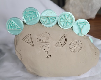 May include: A set of six turquoise blue polymer clay stamps featuring various food and drink designs, including pizza slices, cheese wedges, lime wedges, and a margarita glass. The stamps are arranged in a row on a light brown clay surface.