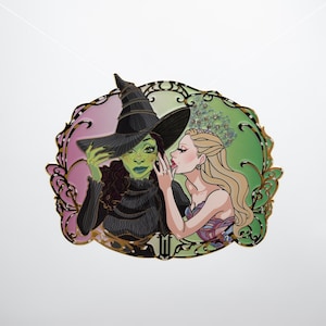 Puede incluir: Pin decorativo con dos mujeres en una ilustración estilizada. Una mujer tiene la piel verde y lleva un sombrero de bruja negro. La otra mujer es rubia y está susurrando. El pin tiene un borde dorado con fondos rosa y verde.