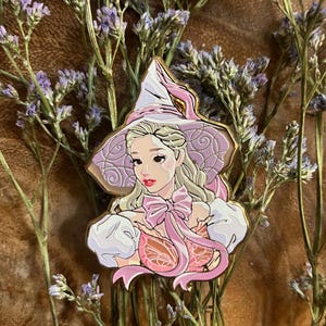 Puede incluir: Un pin de esmalte detallado que representa a una mujer rubia con un sombrero y un vestido de bruja rosa. El pin tiene un contorno dorado y está rodeado de flores moradas. La mujer tiene labios rojos y un lazo rosa.