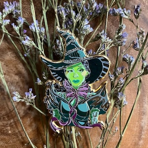 Puede incluir: Un pin de esmalte verde y dorado con una bruja estilizada de piel verde, que lleva un sombrero negro y un lazo morado. El pin está colocado sobre un fondo de flores secas.