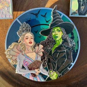 Puede incluir: Un pin esmaltado circular que muestra dos figuras: una persona rubia con un vestido azul claro y rosa con corona, y una persona de piel verde con un vestido negro y sombrero puntiagudo. El fondo muestra un cielo azul verdoso oscuro con siluetas.