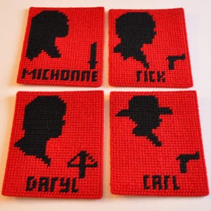 May include: Four red coasters with black silhouettes and names: Michonne, Rick, Daryl, and Carl. Each coaster features a different design, including a knife, a gun, and the number 4. The coasters are made of a woven material.