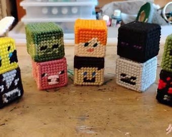 May include: A collection of handmade, pixelated cube-shaped figures, each featuring a different character design. The figures are stacked, showcasing a variety of colors including yellow, green, orange, black, and white. The figures appear to be made of yarn.