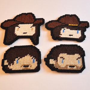 May include: Four handmade felt pixel art coasters featuring characters from the popular TV show The Walking Dead. The coasters are brown, black and white with blue eyes.