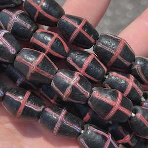 Puede incluir: Hilo de cuentas negras en forma de barril con diseños de cruces rojas y blancas. Las cuentas están ensartadas en un hilo blanco. Las cuentas son de color negro oscuro y mate.