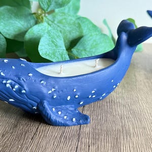 May include: Two blue whale-shaped candles with white speckles. The candles have three wicks and are made of soy wax.