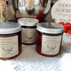 以下が含まれることがあります： シルバーの蓋が付いた茶色のガラス瓶3つに、「Mixology Candle Co. Natural Oven Cleaner with Pure Essential Oils」のラベルが貼られています。瓶は白とグレーのテクスチャのある表面の上に置かれています。